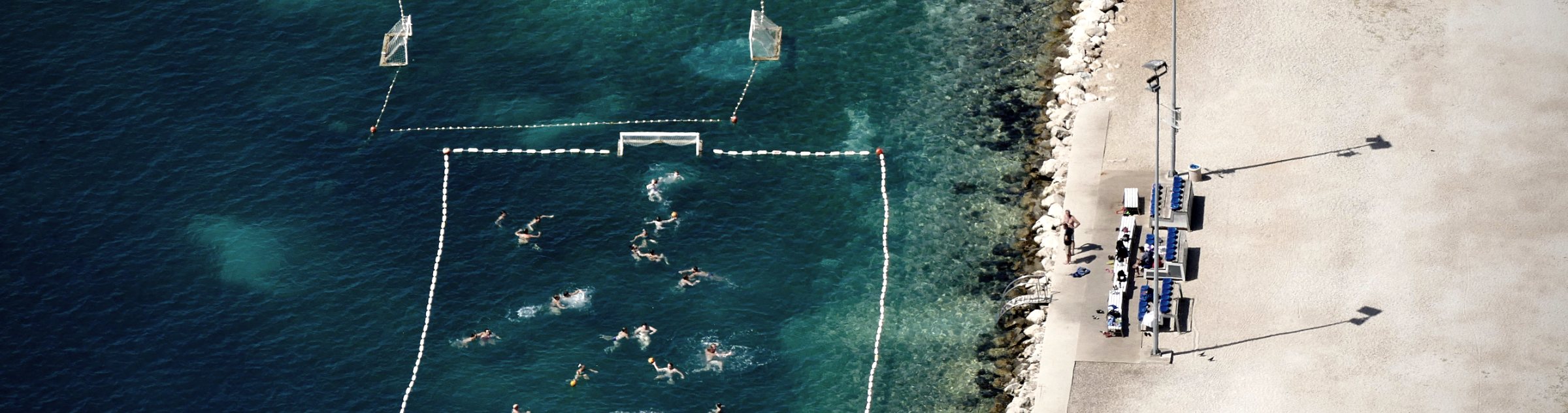 Aerial view of a water polo match played in open water beside a stone shore